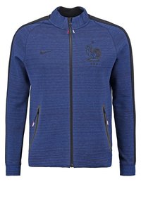 Giacca a zip Nike in tessuto blu con un motivo testurizzato, dotata di pannelli laterali neri, due tasche con zip e un emblema di gallo sul petto.