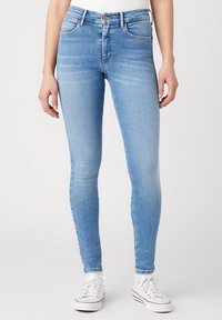 Jeans ajustados de color azul claro en tejido de mezclilla con una textura suave, diseño de cinco bolsillos y pasadores para cinturón, combinados con zapatillas blancas.
