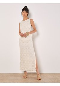 Robe longue sans manches en dentelle crème avec des motifs hexagonaux, dotée d'une fente sur le côté. Des sandales dorées à lanières et des bracelets complètent le look.