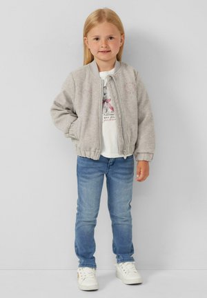 Veste bomber grise avec col et poignets côtelés, t-shirt blanc avec une impression graphique, jeans bleu et baskets blanches. Tissu lisse.