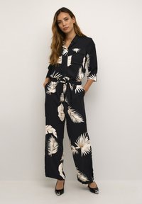 Combinaison noire avec un motif de feuilles de palmier crème, manches longues, patte de boutonnage à l'avant et taille ajustable avec cordon. Des talons noirs brillants complètent le look.