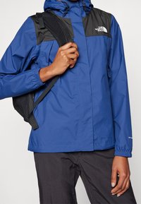 Veste imperméable bleue avec des accents noirs sur les épaules, capuche ajustable, poignets élastiques et logo sur la poitrine ; accompagnée d'un pantalon foncé.