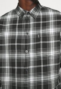 Camicia a quadri bianca e nera con colletto button-down, dotata di taschino sul petto, branding sottile e texture del tessuto visibile nel motivo.