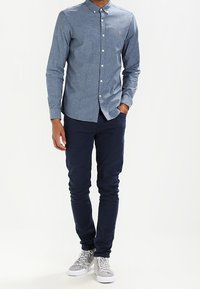 Mężczyzna w niebieskiej koszuli zapinanej na guziki, granatowych dopasowanych spodniach i szarych butach sportowych typu high-top, stojący z jedną ręką w kieszeni.