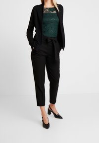 Svart kardiagan över en grön spetsbodysuit, kombinerad med svarta skräddarsydda byxor och svarta klackskor. Elegant, strukturerad silhuett.