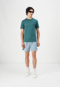 Türkiser Baumwoll-T-Shirt mit kurzen Ärmeln, kombiniert mit hellblauen Shorts. Das Outfit wird mit weißen Sneakers und einer schwarzen Sonnenbrille abgerundet.