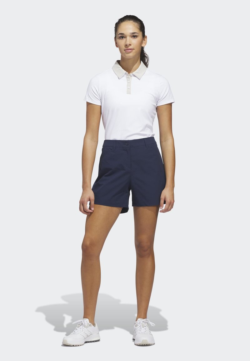 Νεαρή γυναίκα που στέκεται φορώντας λευκό πόλο πουκάμισο, σκούρα μπλε σορτς και λευκά αθλητικά παπούτσια σε απλό φωτεινό φόντο.