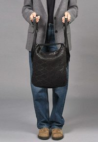 Zwarte leren handtas met een geweven patroon, voorzien van een enkele schouderband en een zachte, gestructureerde vorm. Draagt door een persoon in casual kleding.