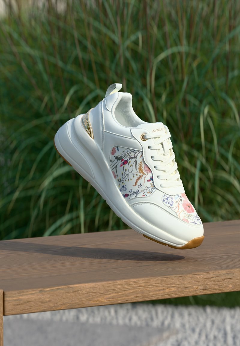 Λευκό αθλητικό παπούτσι με λουλουδάτο σχέδιο και χρυσές λεπτομέρειες που αιωρείται πάνω από ένα ξύλινο παγκάκι με φόντο πράσινο γρασίδι.