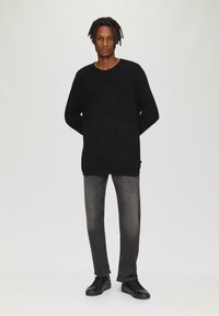Schwarzer Strickpullover mit rundem Halsausschnitt, kombiniert mit dunkelgrauen Jeans und schwarzen Turnschuhen. Gerader Schnitt, minimalistische Details und strukturierter Stoff.