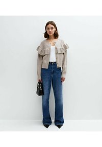 Beige kofta med volangdetaljer vid axlarna, guldknappar, i kombination med blå utsvängda jeans och en liten svart handväska.