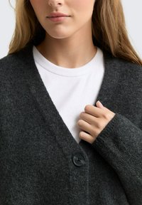 Temno siva cardigan iz mehke tkanine, s popustom v obliki v, dolgimi rokavi in enim gumbom za zapiranje. Pod njim je bela srajca.