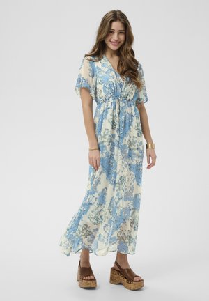 Femme aux longs cheveux bruns portant une longue robe fleurie claire avec des motifs bleus et des sandales à plateforme marron, souriante et debout sur un fond uni.