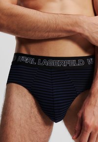 Kratke hlače u tamnoplavoj i crnoj prugastoj kombinaciji s širokim elastičnim pojasom na kojem je bijeli logotip "KARL LAGERFELD". Mekani, prilagodljivi materijal.