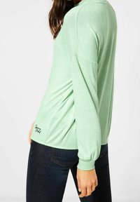 Košile s dlouhým rukávem v mintově zelené barvě, s lehkou strukturou, uvolněným střihem a logem blízko dolního lemu. Kombinováno s tmavými džíny.
