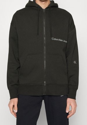 Felpa nera con zip, tasche frontali e logo "Calvin Klein Jeans" sul petto e "cK" sulla manica sinistra, indossata da una persona davanti a uno sfondo semplice.