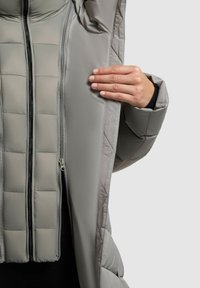 Grå puffjacka med zip-stängning, quiltad textur och en mjuk innerfoder. Hand placerad nära öppningen för detalj.