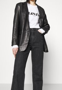 Schwarze Lederblazer über einem weißen Grafik-T-Shirt mit dem Text "Levi's", kombiniert mit dunkelblauen, weiten Denim-Jeans.
