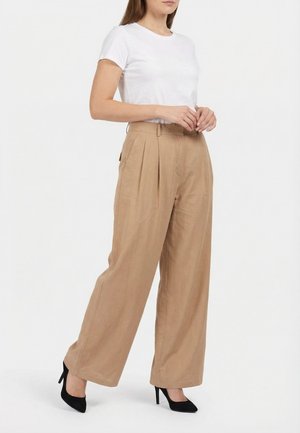 Femme portant un pantalon large beige, un haut blanc à manches courtes, et des chaussures à talons noirs, debout devant un fond blanc.