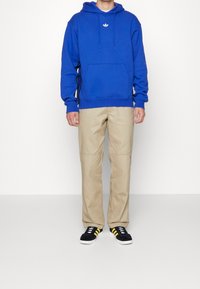 Sudadera azul con bolsillo frontal y cordones, pantalones caqui con parches en las rodillas y zapatillas negras con rayas amarillas.