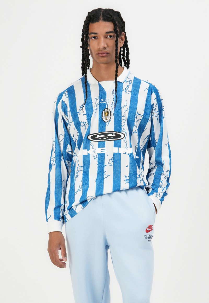 Camisa de manga larga con rayas verticales azules y blancas, que presenta un patrón texturizado, cuello y un logotipo central. Combinada con pantalones de chándal azul claro.