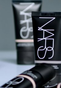 Tubes cosmétiques noirs avec des lettres blanches. Un récipient est positionné verticalement ; l'autre est légèrement incliné. Le texte comprend "NARS" et des détails sur le produit.
