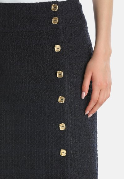 Close-up of dark textured skirt with a vertical row of seven decorative square gold buttons and a hand with manicured nails resting beside it.