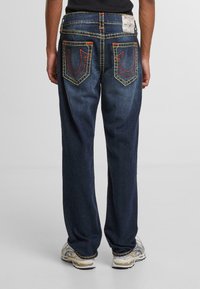 Jeans de mezclilla azul con corte recto, que presentan costuras amarillas, diseños de bolsillos traseros en contraste y un parche de marca en la cintura.