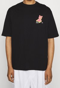 Mustat puuvillateepaita, jossa on värikäs karhukuva vasemmassa rinnassa, pyöreä pääntie ja lyhyet hihat, löysä istuvuus.