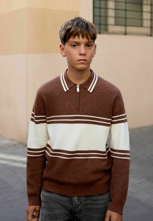 Teenagedreng står udendørs iført en brun og hvid stribet pullover med lynlås ved kraven og grå bukser.