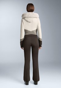 Női sídzseki krém színben, műszőrme kapucnival és szürke steppelt részletekkel; barna, testhezálló sí nadrággal párosítva. Sima textúra és szabott illeszkedés.