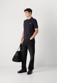 Ναυτικό μπλουζάκι πόλο με κοντά μανίκια, λογότυπο στο στήθος, συνδυασμένο με μαύρα τζιν κατευθείαν ποδιών και μαύρα αθλητικά παπούτσια, κρατώντας μια μαύρη τσάντα ώμου.