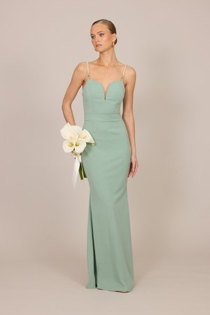 Frau in bodenlangem salbeigrünem Kleid mit Perlen-Trägern, die weiße Calla-Lilien vor einem schlichten beigen Hintergrund hält.