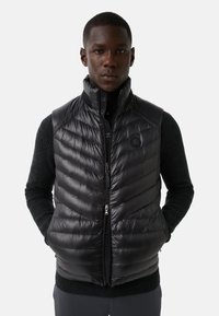 Zwarte pufferbodywarmer met een hoge kraag, chevron-gestikt ontwerp en een rits aan de voorkant. Heeft een logodetail op de borst en is gemaakt van glanzend materiaal.