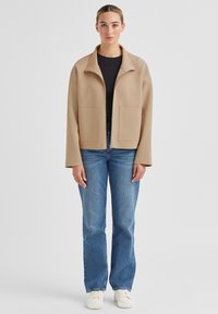 Beige Reißverschlussjacke mit hohem Kragen, Fronttaschen und lockerem Schnitt, kombiniert mit blauen Jeans und weißen Turnschuhen.