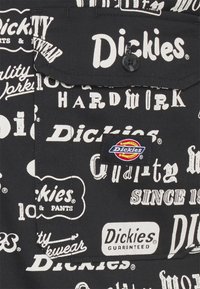 Tissu noir avec logos et motifs de texte blancs de la marque Dickies, avec une poche équipée d’un bouton Dickies et d’un patch logo cousu.