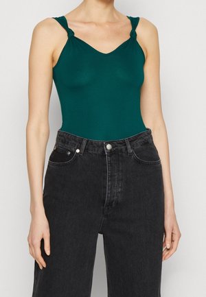 Femme portant un haut sans manches vert foncé avec des bretelles nouées, rentré dans un jean noir délavé taille haute.