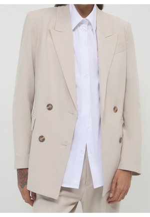 Blazer beige à double boutonnage avec des boutons en écaille porté sur une chemise blanche boutonnée et un pantalon beige assorti.