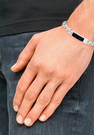 Mannelijke hand met een zilveren kettingarmband met een zwarte marmerinleg die rust op de zak van een donkerdenim broek.
