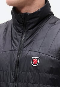 Inimene musta tikitud jopega, millel on kõrge krae ja punane rebaste logo rinnal.