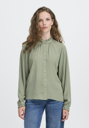 Woman with long wavy hair wears a light green buttoned blouse with ruffled collar and cuffs, paired with blue jeans, standing against white background.