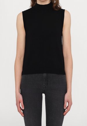 Mujer que lleva una camiseta negra sin mangas con cuello alto simulado y vaqueros ajustados de color gris oscuro, de pie con los brazos a los lados sobre un fondo liso.