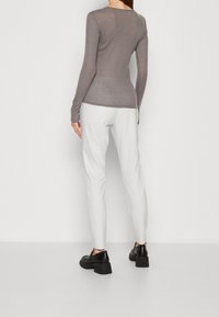 Person som bär en figurnära grå långärmad topp, vita smala byxor och svarta plattformsträskor, som står mot en enkel vit bakgrund.