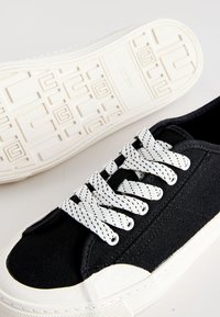 Baskets en toile noire avec semelle en caoutchouc blanche et embout blanc, lacets blancs à pois noirs, et motif texturé de la semelle visible en dessous.