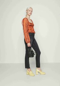 Blusa laranja com padrão de cobra, com mangas de balão, jeans skinny pretos, sapatos de salto bloco amarelos e uma pequena mala preta.