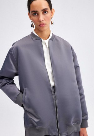 Jachetă bomber gri din satin cu guler înalt, deschidere cu fermoar, buzunare laterale și accente ribbed la manșete și la tiv. Textură netedă.