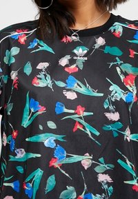 Czarny t-shirt w kwiatowy wzór z mieszanką niebieskich, czerwonych i różowych kwiatów oraz zielonych liści. Logo Adidas umieszczone na środku klatki piersiowej. Miękka, gładka tkanina.