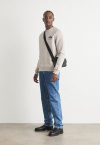 Beige sweatshirt med logo, blå lige ben jeans og sorte læderstøvler. Sort crossover taske hviler på venstre skulder. Ensfarvet baggrund.