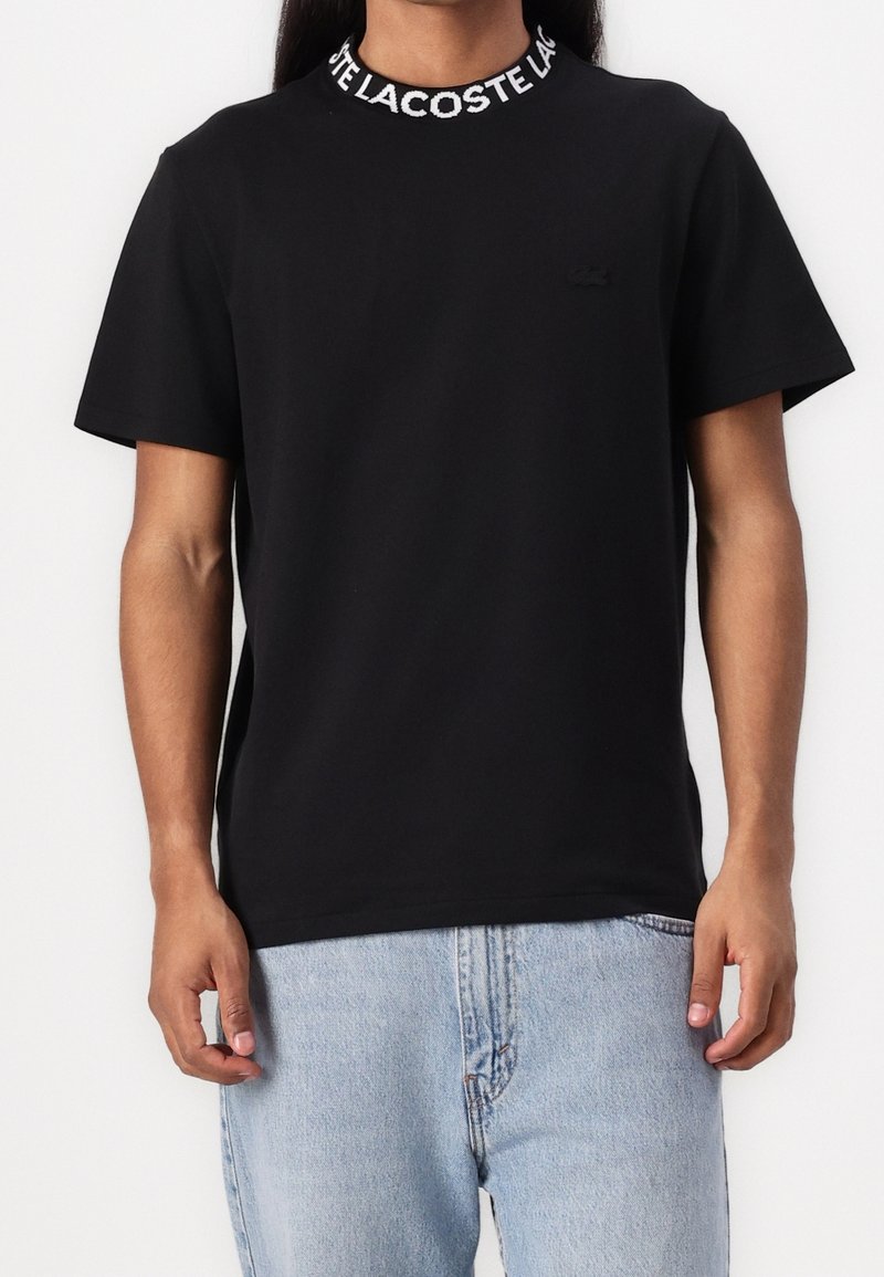 Personne portant un t-shirt noir à manches courtes avec le texte "LACOSTE" sur le col et un jean bleu clair.