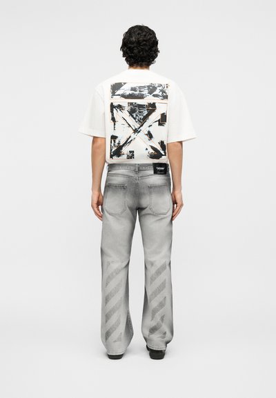 Άτομο με σγουρά μαλλιά στέκεται γυρισμένο προς τα πίσω, φοράει λευκό μπλουζάκι με μαύρα γραφικά βέλη και άνετο γκρι τζιν με διαγώνια ριγέ σχέδια.